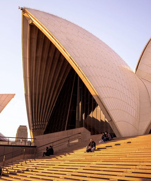 Sydney Opera House at sunset, featuring its iconic shell-like structures and people sitting on the steps in the foreground.