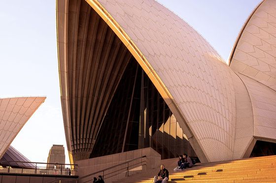 Sydney Opera House at sunset, featuring its iconic shell-like structures and people sitting on the steps in the foreground.