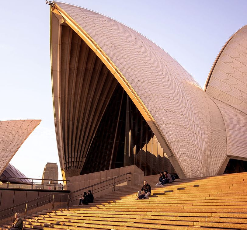 Sydney Opera House at sunset, featuring its iconic shell-like structures and people sitting on the steps in the foreground.