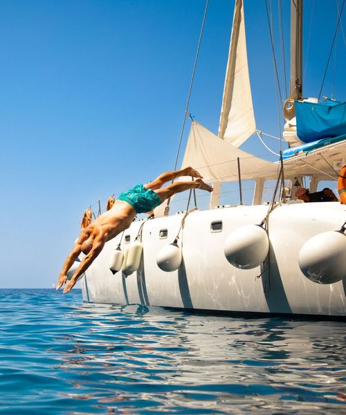 A person in swimwear dives off the side of a white sailboat into the blue ocean under a clear sky.