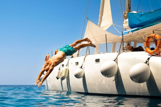A person in swimwear dives off the side of a white sailboat into the blue ocean under a clear sky.