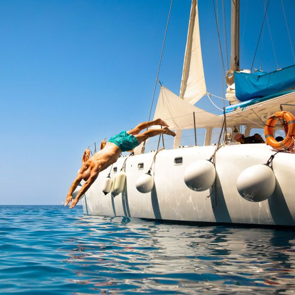 A person in swimwear dives off the side of a white sailboat into the blue ocean under a clear sky.