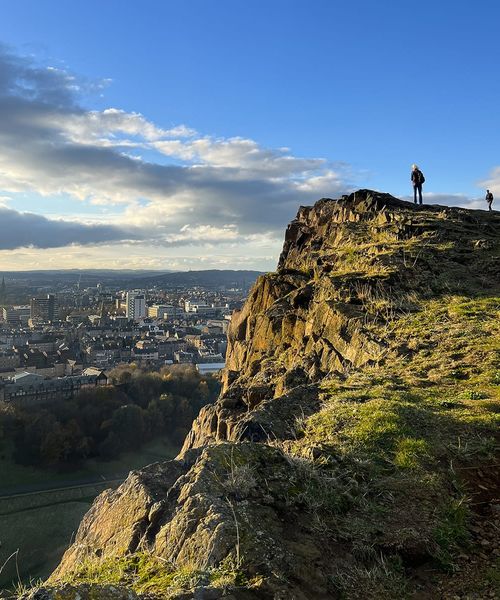 Person standing on a rocky hilltop overlooking a cityscape under a clear blue sky with scattered clouds.