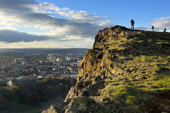 Person standing on a rocky hilltop overlooking a cityscape under a clear blue sky with scattered clouds.