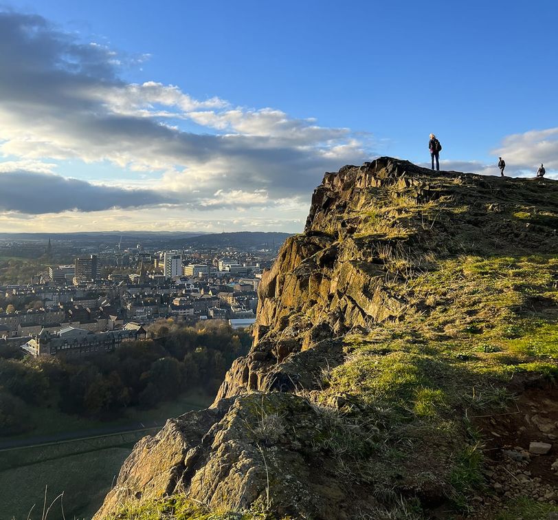 A rocky cliff overlooks a cityscape under a blue sky with clouds. People stand on the edge, admiring the view.