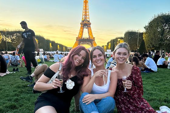 Three women sit on grass, smiling and holding drinks, with the Eiffel Tower illuminated in the background at sunset.