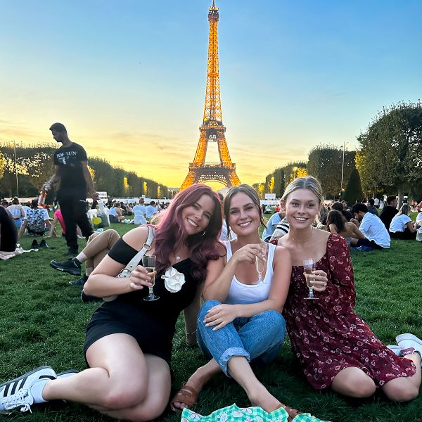 Three women sit on grass, smiling and holding drinks, with the Eiffel Tower illuminated in the background at sunset.