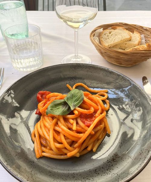 A plate of pasta with tomato sauce and basil on a gray plate, alongside bread in a basket and a glass of white wine on a white tablecloth.