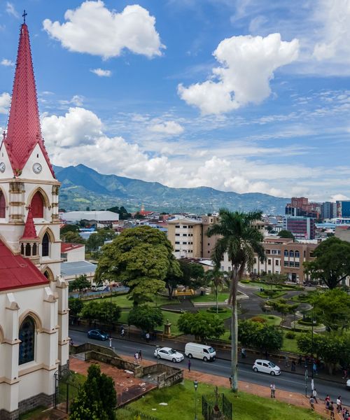 A cityscape with a church featuring a red steeple in the foreground, lush green trees, roads, and mountains in the background under a cloudy sky.