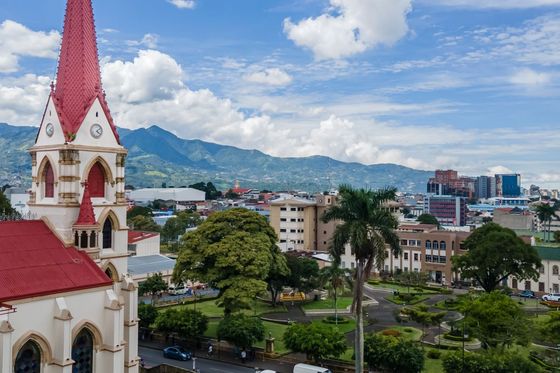 A cityscape with a church featuring a red steeple in the foreground, lush green trees, roads, and mountains in the background under a cloudy sky.