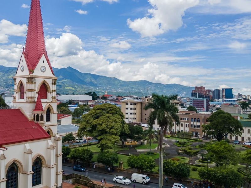 A cityscape with a church featuring a red steeple in the foreground, lush green trees, roads, and mountains in the background under a cloudy sky.