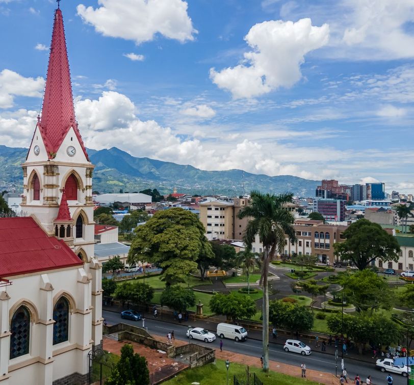 A cityscape with a church featuring a red steeple in the foreground, lush green trees, roads, and mountains in the background under a cloudy sky.
