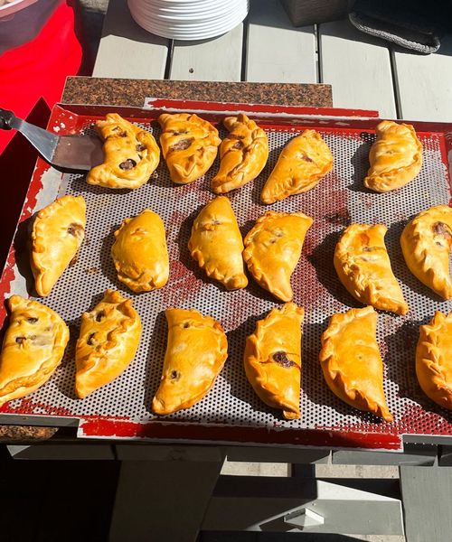 A tray of golden-brown empanadas on a perforated baking sheet, with a person removing one with a spatula.
