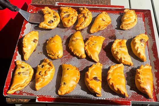A tray of golden-brown empanadas on a perforated baking sheet, with a person removing one with a spatula.