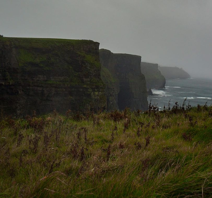 Dramatic cliffs under a cloudy sky, overlooking a turbulent sea, with lush green grass in the foreground.
