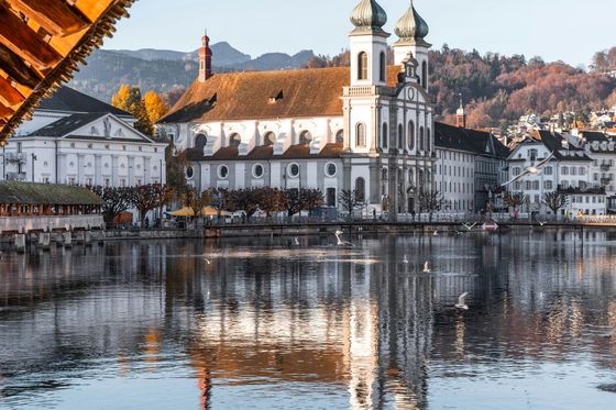 Riverside view of a baroque church with twin towers, reflecting in the water, surrounded by autumn trees and mountains in the backdrop.