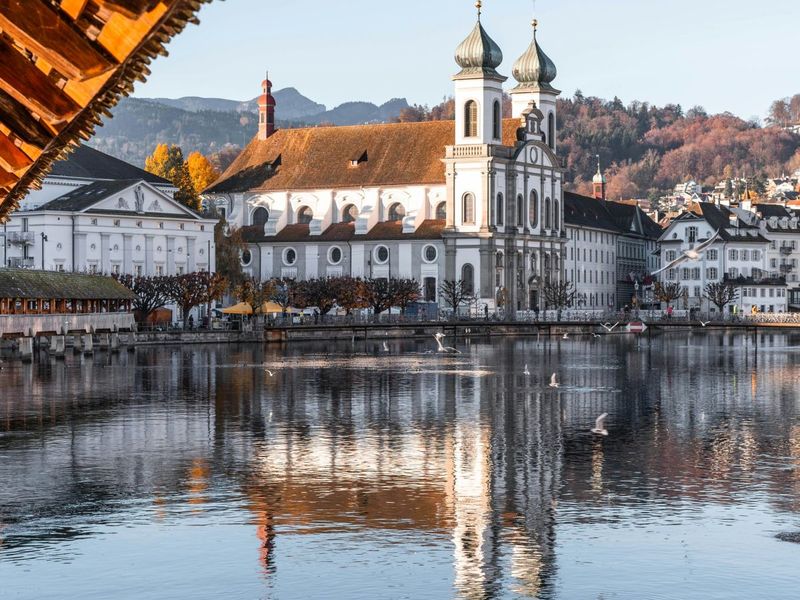 Riverside view of a baroque church with twin towers, reflecting in the water, surrounded by autumn trees and mountains in the backdrop.