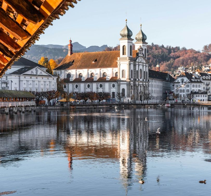 Riverside view of a baroque church with twin towers, reflecting in the water, surrounded by autumn trees and mountains in the backdrop.