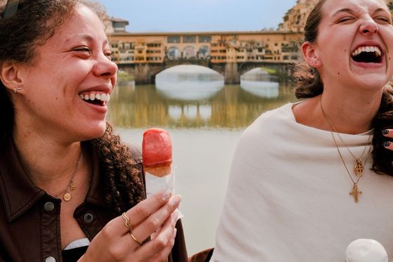 Two women laughing and enjoying ice cream cones by a river, with a bridge in the background.