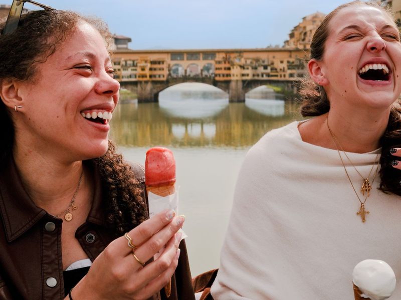 Two women laughing and enjoying ice cream cones by a river, with a bridge in the background.