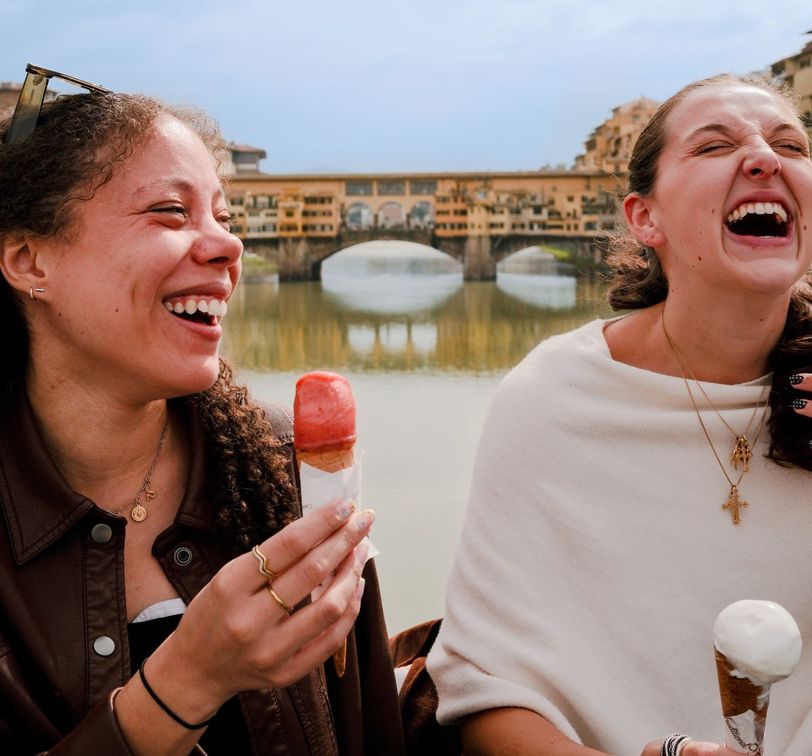 Two women laughing and enjoying ice cream cones by a river, with a bridge in the background.