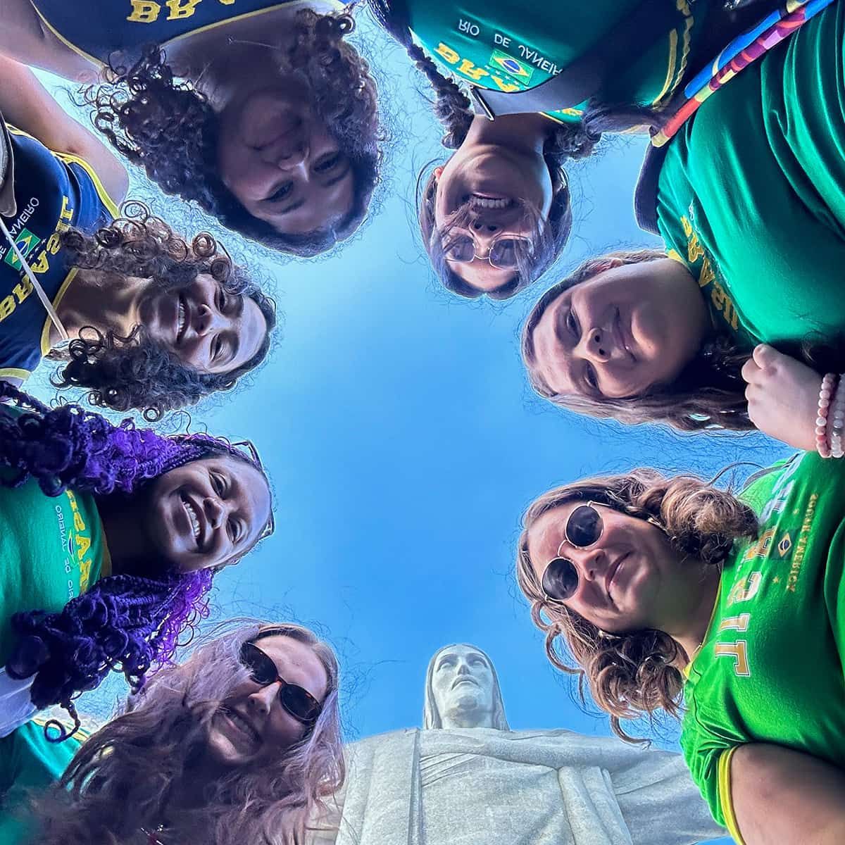 A group of people in a circle, smiling and looking down at the camera, with the Christ the Redeemer statue visible in the background.