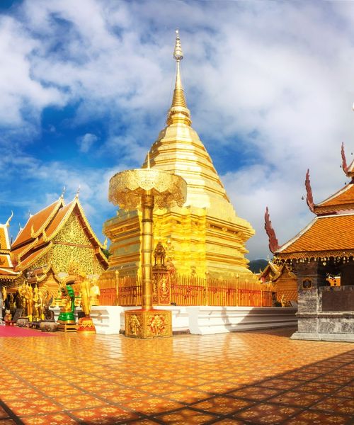 A golden Buddhist temple with intricate rooftops and a large golden stupa set against a blue sky with scattered clouds.