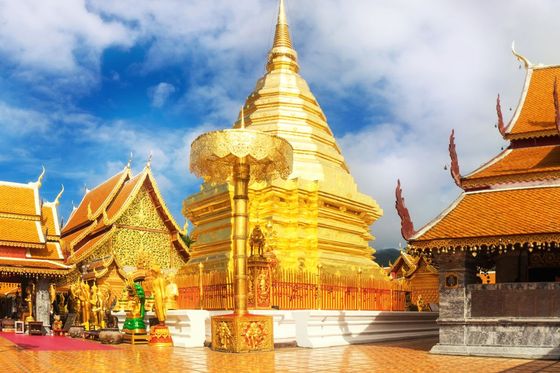 A golden Buddhist temple with intricate rooftops and a large golden stupa set against a blue sky with scattered clouds.