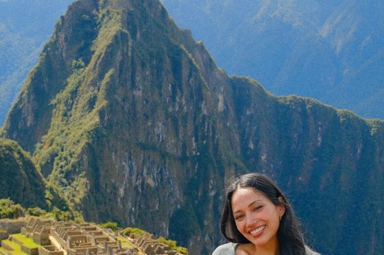 A woman smiling in front of the ancient stone ruins of Machu Picchu, with a large, lush mountain in the background.