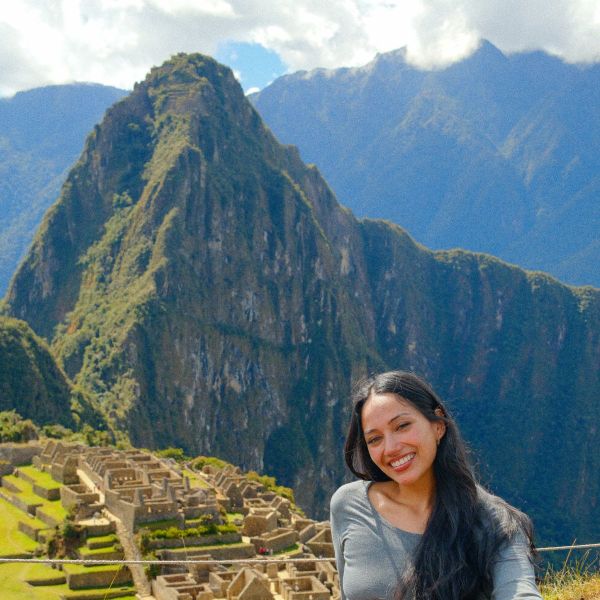 A woman smiling in front of the ancient stone ruins of Machu Picchu, with a large, lush mountain in the background.