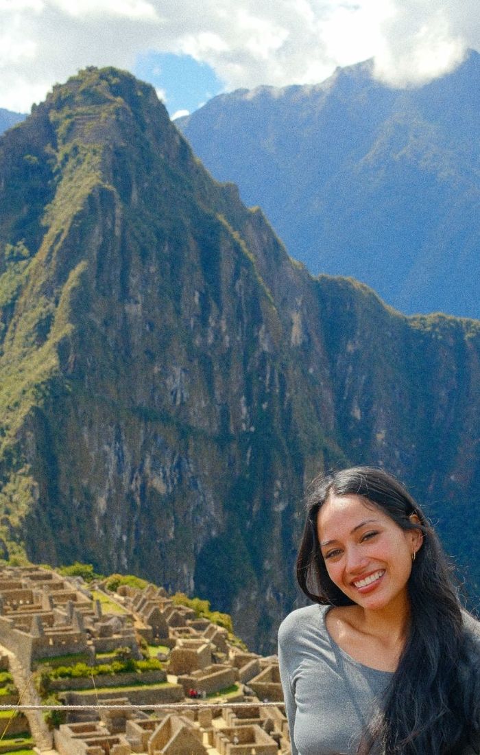 A woman smiling in front of the ancient stone ruins of Machu Picchu, with a large, lush mountain in the background.
