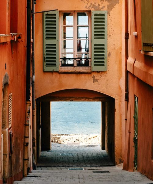 Narrow alley with orange buildings and green shutters leading to a bright view of the sea under an archway.