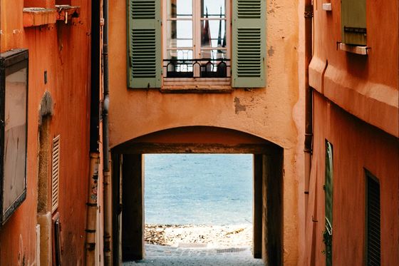 Narrow alley with orange buildings and green shutters leading to a bright view of the sea under an archway.