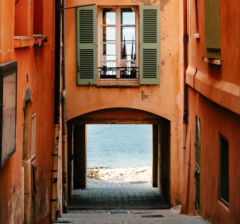 Narrow alley with orange buildings and green shutters leading to a bright view of the sea under an archway.