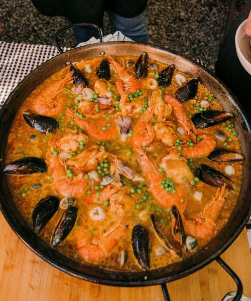 A pan of seafood paella with prawns, mussels, and peas on a wooden board, next to a glass of red wine.