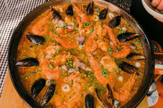 A pan of seafood paella with prawns, mussels, and peas on a wooden board, next to a glass of red wine.