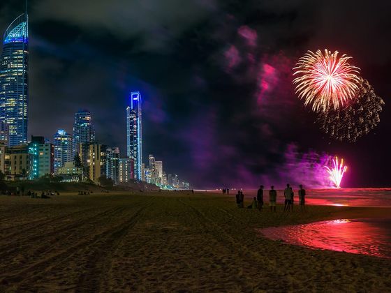 Fireworks light up the night sky over a city beach. Skyscrapers are illuminated, and silhouettes of people watch the display near the shoreline.