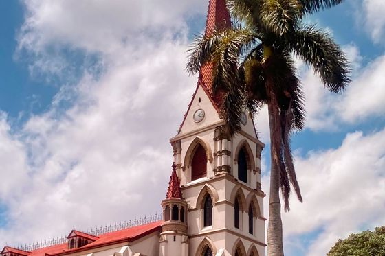 A historic church with a red-roofed steeple stands beside a tall palm tree under a partly cloudy sky.