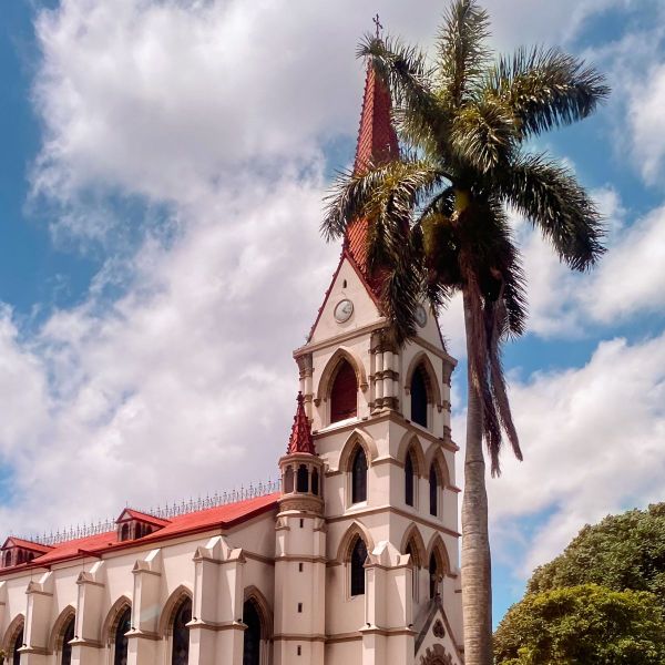 A historic church with a red-roofed steeple stands beside a tall palm tree under a partly cloudy sky.