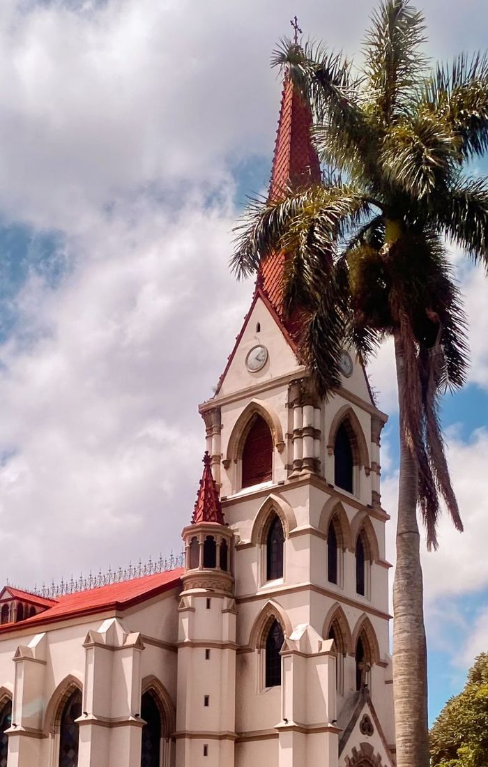 A historic church with a red-roofed steeple stands beside a tall palm tree under a partly cloudy sky.
