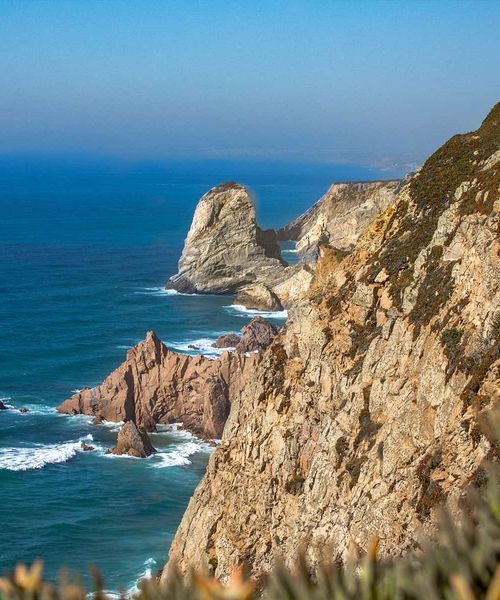 Rocky coastline with steep cliffs overlooking a deep blue ocean under a clear sky, featuring a prominent rock formation in the distance.