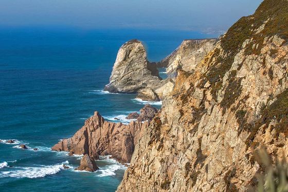 Rocky coastline with steep cliffs overlooking a deep blue ocean under a clear sky, featuring a prominent rock formation in the distance.