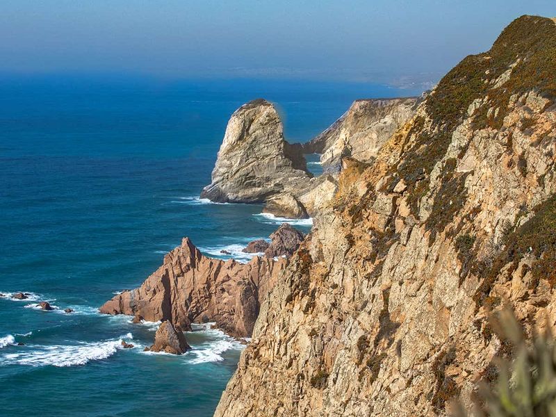 Rocky coastline with steep cliffs overlooking a deep blue ocean under a clear sky, featuring a prominent rock formation in the distance.