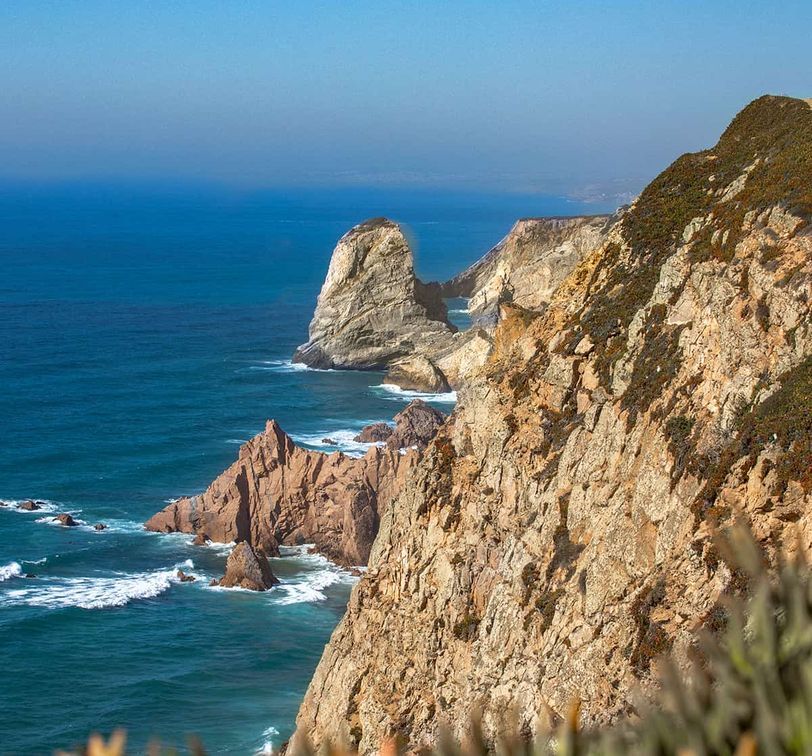 Rocky coastline with steep cliffs overlooking a deep blue ocean under a clear sky, featuring a prominent rock formation in the distance.
