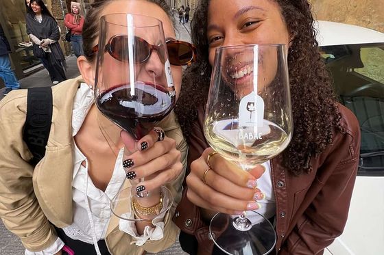 Two women smiling and holding oversized wine glasses on a street with arched buildings in the background.