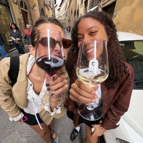 Two women smiling and holding oversized wine glasses on a street with arched buildings in the background.