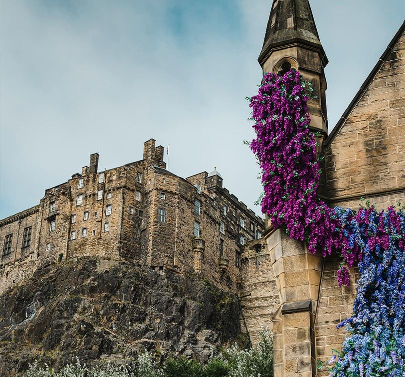 Historic stone castle on a rocky hill with a foreground of vibrant purple and blue flowers cascading down a chapel wall.