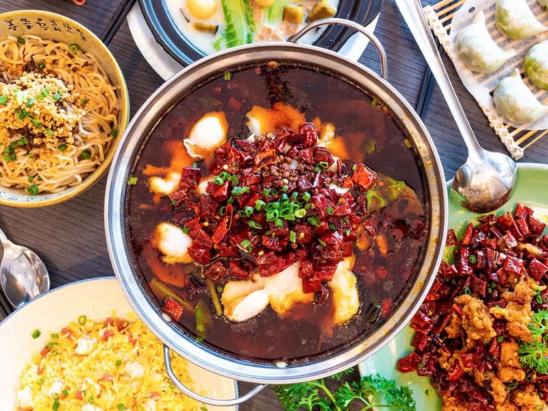 A vibrant spread of Asian dishes features a spicy hot pot, stir-fried noodles, dumplings, and colorful rice on a wooden table.
