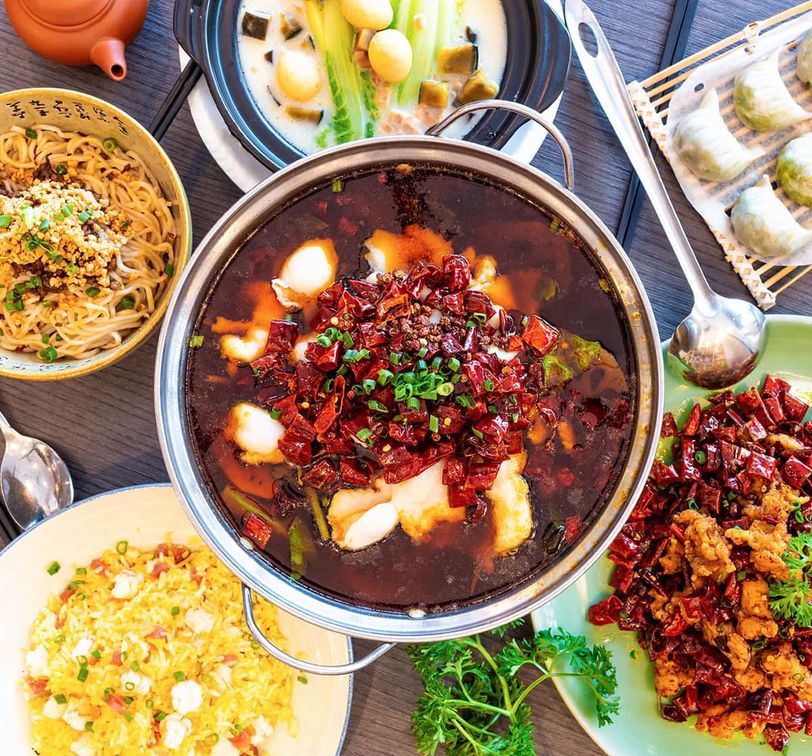 A vibrant spread of Asian dishes features a spicy hot pot, stir-fried noodles, dumplings, and colorful rice on a wooden table.