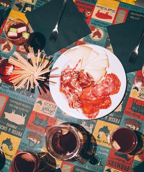 A table with a plate of cured meats and cheese, surrounded by drinks, napkins, and forks on a colorful, patterned tablecloth.
